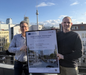 Zwei Männer stehen auf einem Dach und halten ein gerahmtes Plakat über ein Berliner Zukunftsprojekt in den Händen; im Hintergrund sind Gebäude der Stadt und der Berliner Fernsehturm zu sehen.